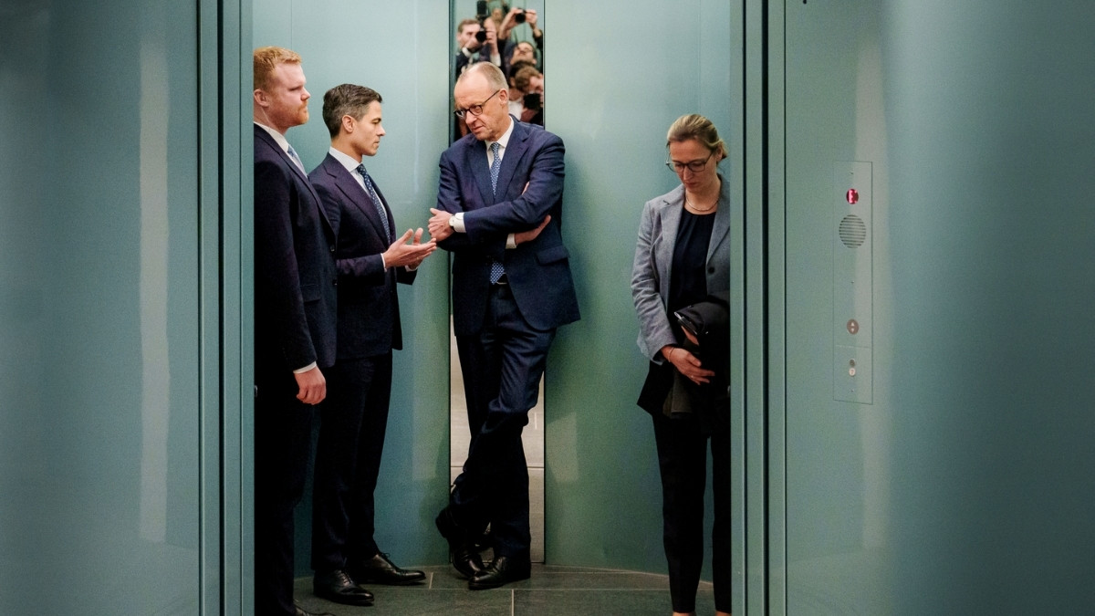 Bundeskanzler Friedrich Merz steht mit weiteren Personen in einem Aufzug und hört einem Gespräch zu