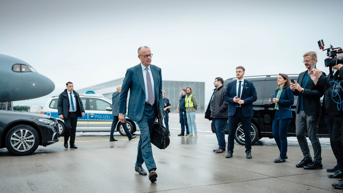 Mann im Anzug geht auf einem Flughafengelände an Fahrzeugen und Kamerateam vorbei