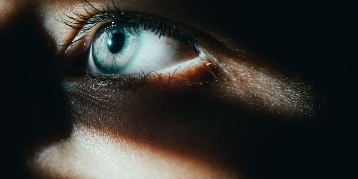 Nahaufnahme eines hellblauen Auges mit deutlich sichtbaren Wimpern und Lichtreflexen, teilweise von Schatten über dem Gesicht umgeben.