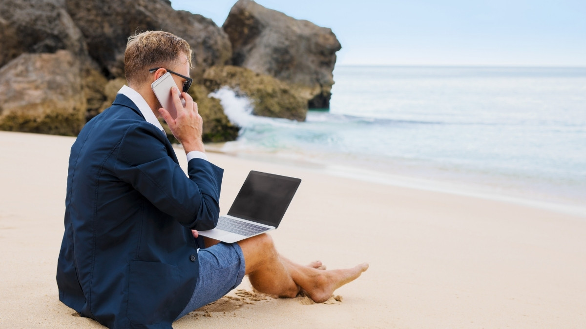 Mann im Anzug telefoniert mit Laptop am Strand vor dem Meer