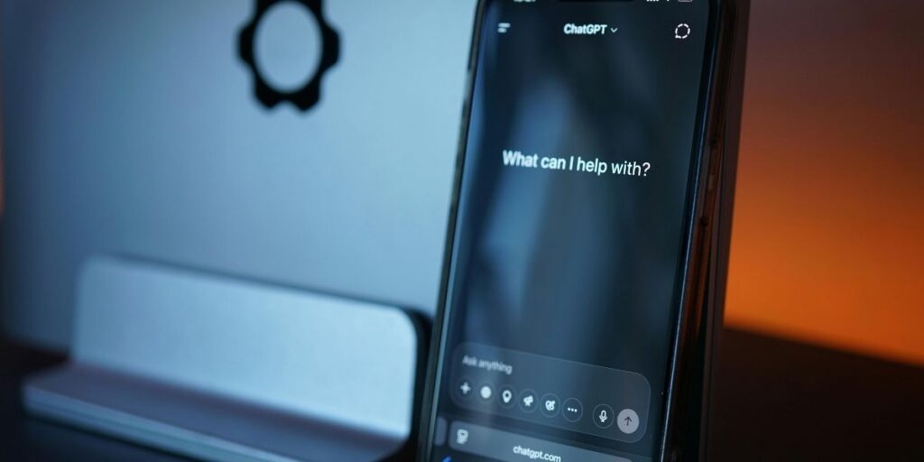 Smartphone mit geöffneter ChatGPT-App vor einem unscharfen Computerbildschirm auf einem Schreibtisch.