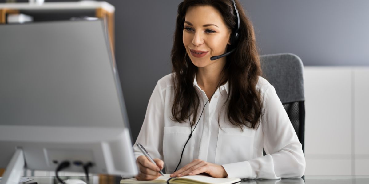 Lächelnde Frau mit Headset sitzt am Schreibtisch, blickt auf einen Monitor und macht sich Notizen.