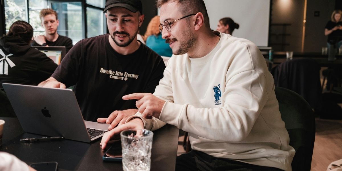 Zwei Personen arbeiten gemeinsam an einem Laptop in einem modernen Büro, weitere Personen sitzen im Hintergrund.