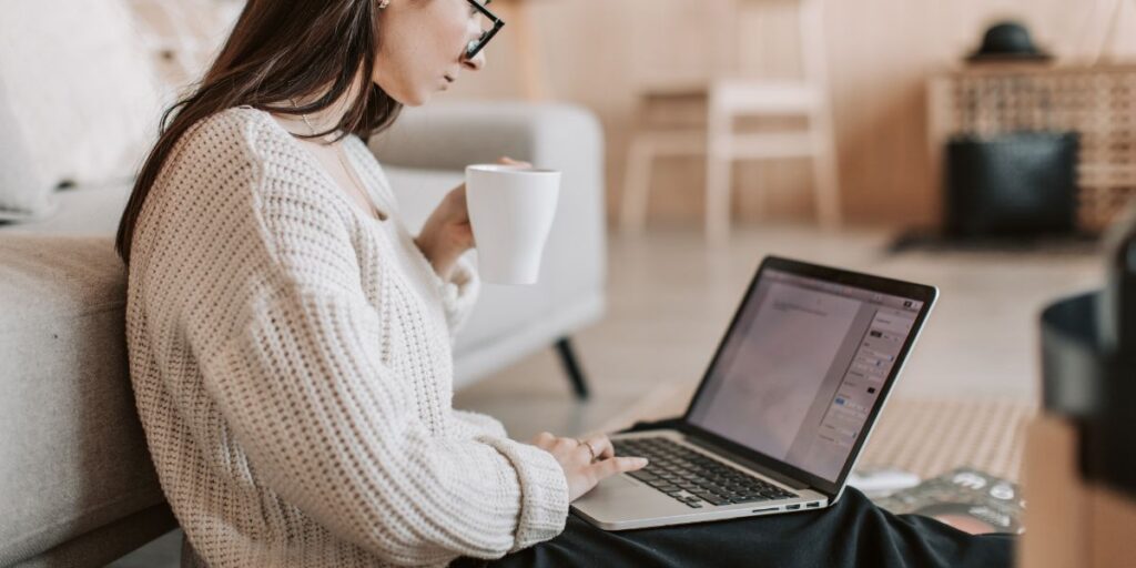Person mit Brille sitzt auf dem Boden neben einem Sofa, hält eine Tasse und arbeitet an einem Laptop im Wohnzimmer.