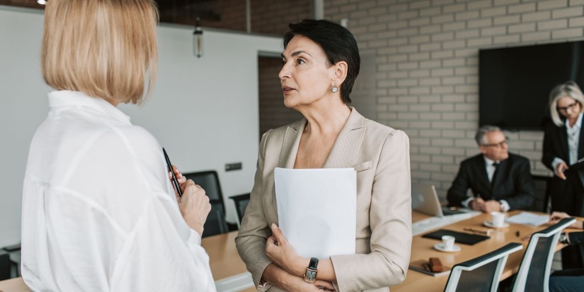 Zwei Personen sprechen in einem Konferenzraum; eine Person hält Dokumente, weitere Personen sitzen im Hintergrund am Besprechungstisch.