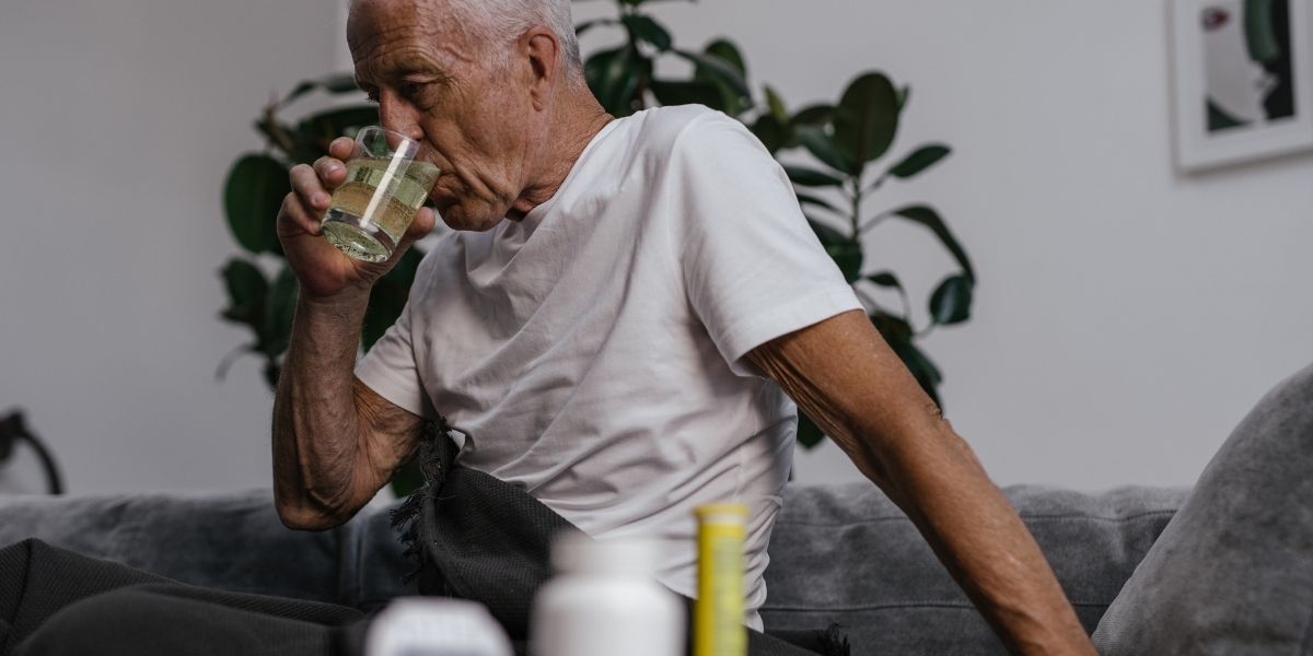Es sitzt ein älterer Mann auf Sofa und trinkt Wasser.