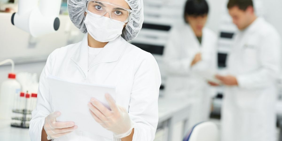 Laborfachkraft mit Schutzmaske, Haube, Schutzbrille und Handschuhen hält ein Tablet in einem Labor; im Hintergrund arbeiten zwei weitere Personen an einem Labortisch.