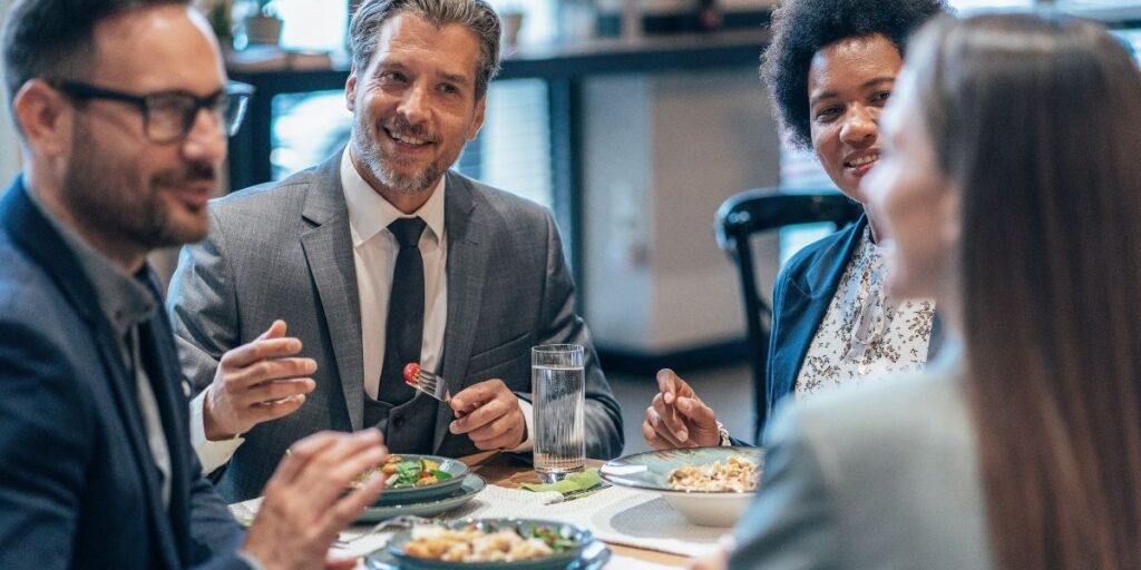 Vier Geschäftsleute sitzen während eines Essens an einem Restauranttisch, lächeln und unterhalten sich in einer entspannten Meeting-Atmosphäre.