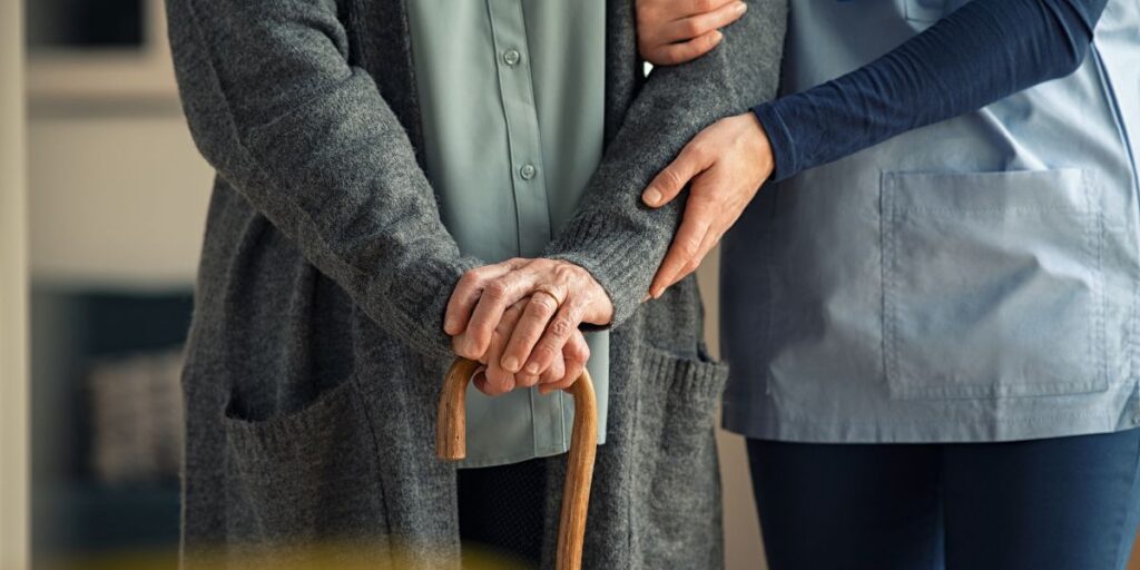 Pflegekraft unterstützt eine ältere Person mit Gehstock, beide stehen nebeneinander in einem Innenraum.