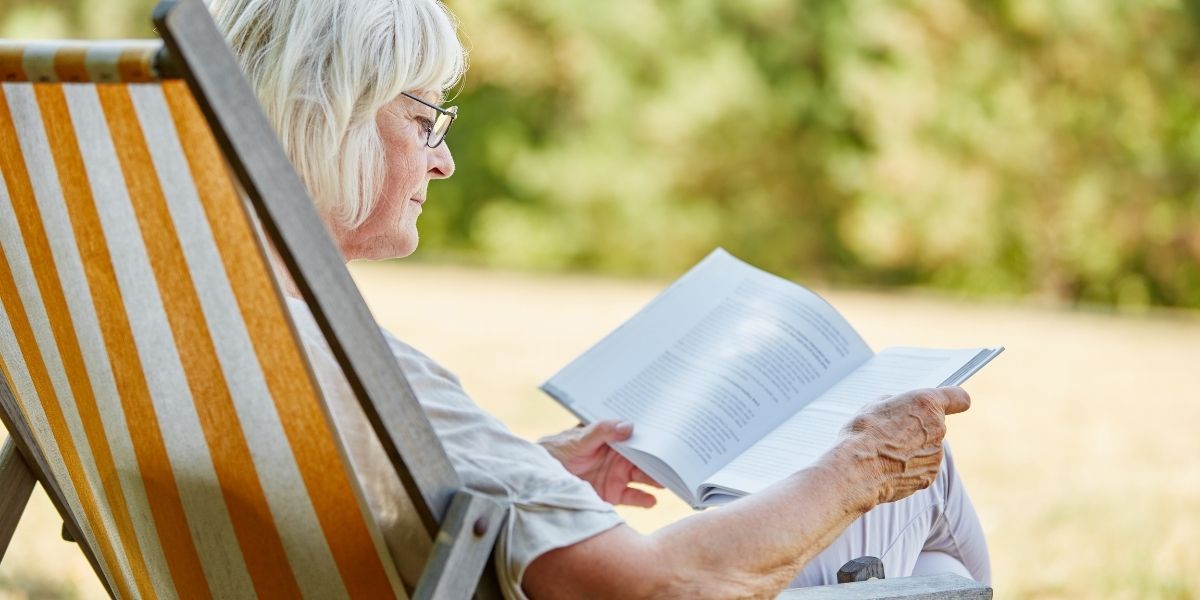 Ältere Person mit Brille liest ein Buch in einem Liegestuhl im Freien.