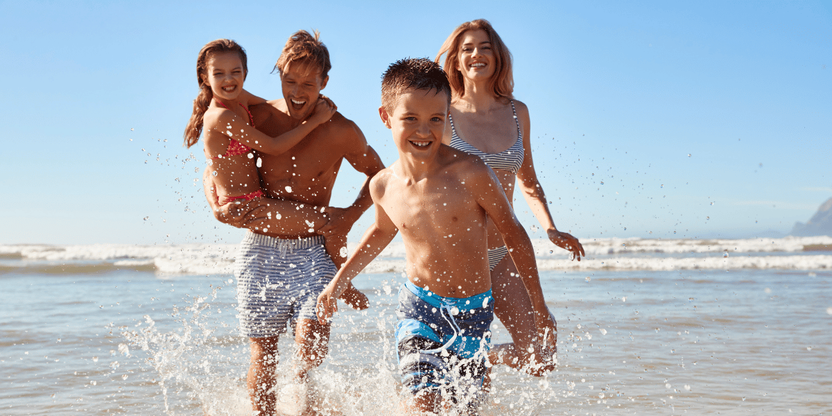 Eine vierköpfige Familie läuft lachend durch das flache Meer an einem sonnigen Strand. Ein Junge rennt voraus und spritzt Wasser auf, während ein Mann ein Mädchen auf dem Arm trägt. Eine Frau folgt ihnen. Blauer Himmel und Wellen unterstreichen die sommerliche Urlaubsstimmung.