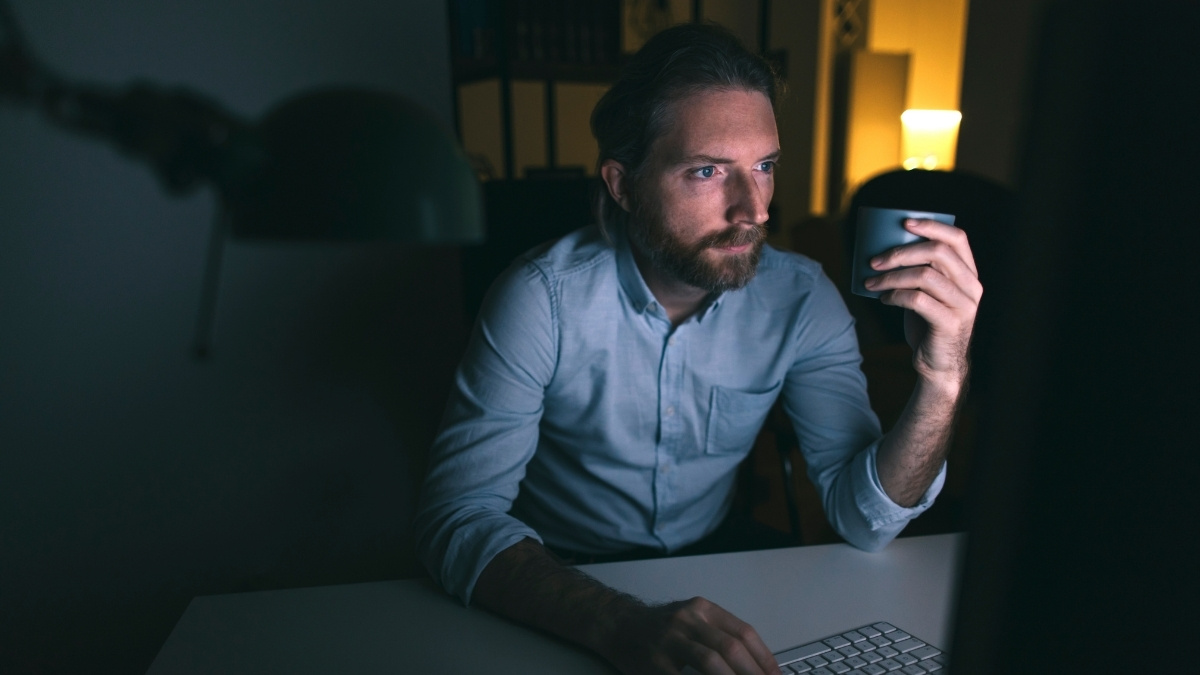 Mann arbeitet nachts am Computer und hält eine Tasse in der Hand