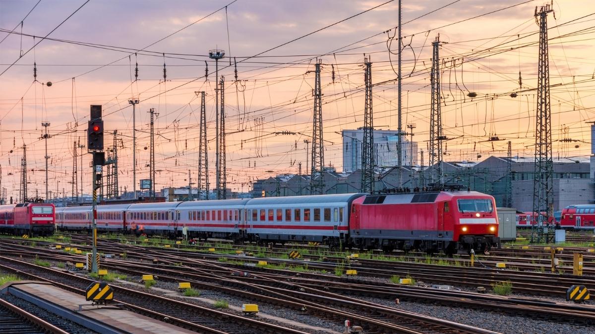 Zug mit roter Lokomotive fährt durch einen großen Bahnhof mit vielen Oberleitungen bei Sonnenuntergang