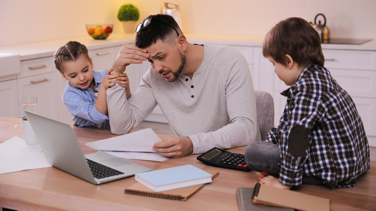 Vater sitzt mit zwei Kindern, Unterlagen und Laptop am Küchentisch
