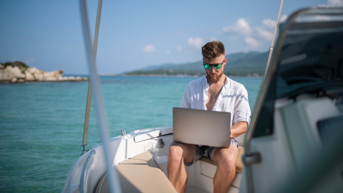 Mann arbeitet mit Laptop auf einem Boot vor türkisblauem Meer