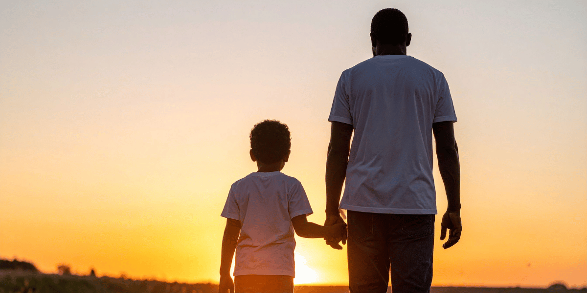 Erwachsener und Kind gehen Hand in Hand einem Sonnenuntergang entgegen. Beide sind von hinten zu sehen und tragen helle T-Shirts – das Bild steht symbolisch für Zukunft, Familie und finanzielle Vorsorge.