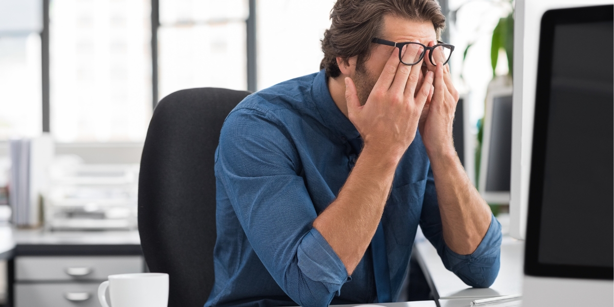 Person mit Brille sitzt an einem Schreibtisch vor einem Computer und reibt sich mit beiden Händen die Augen. Im Vordergrund steht eine Tasse, im Hintergrund sind Bürofenster zu sehen.