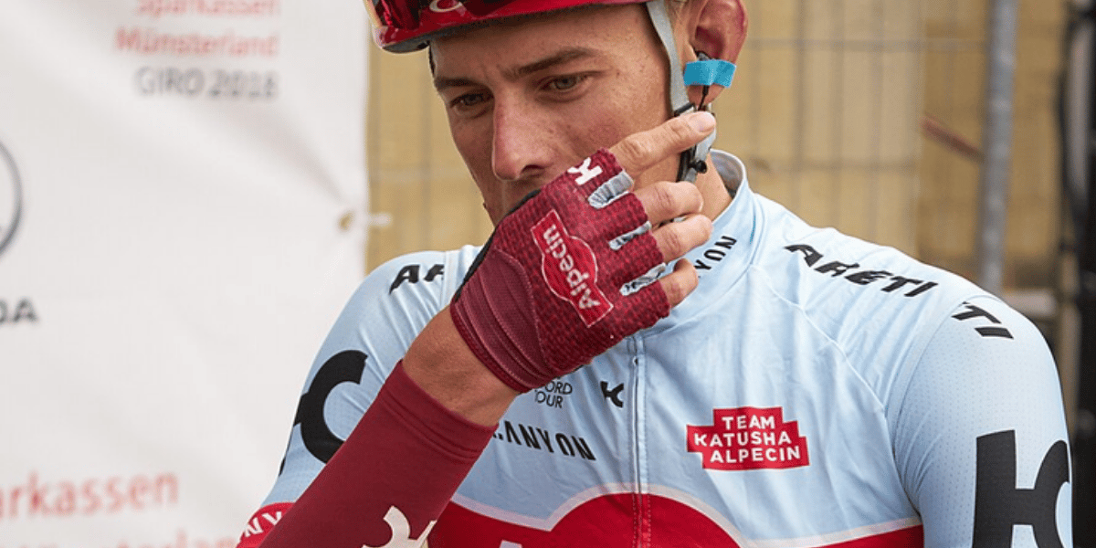 Nahaufnahme eines Rennradfahrers im Trikot und Helm, der mit behandschuhter Hand sein Gesicht berührt.
