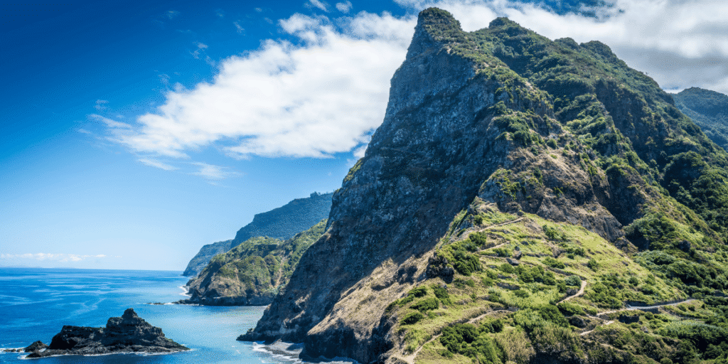 Steile, gr&uuml;n bewachsene Felsk&uuml;ste mit Wanderweg am Meer unter blauem Himmel mit wei&szlig;en Wolken.