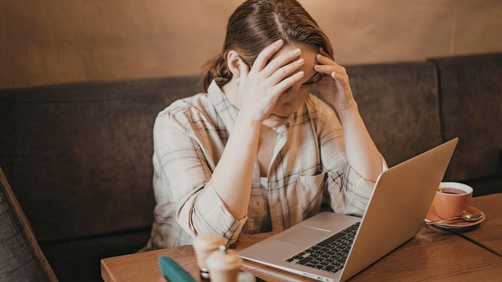 Frau sitzt am Tisch vor einem Laptop und h&auml;lt sich ersch&ouml;pft den Kopf.