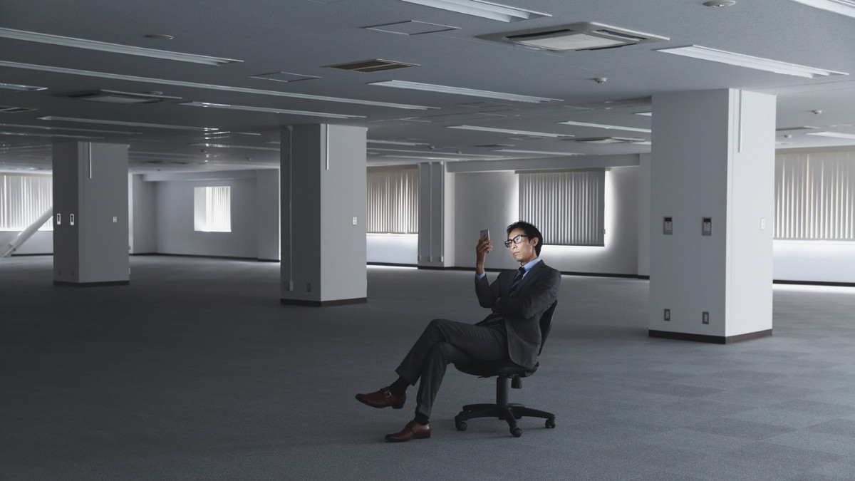 Mann im Anzug sitzt allein in einem leeren Büro und schaut auf sein Smartphone.