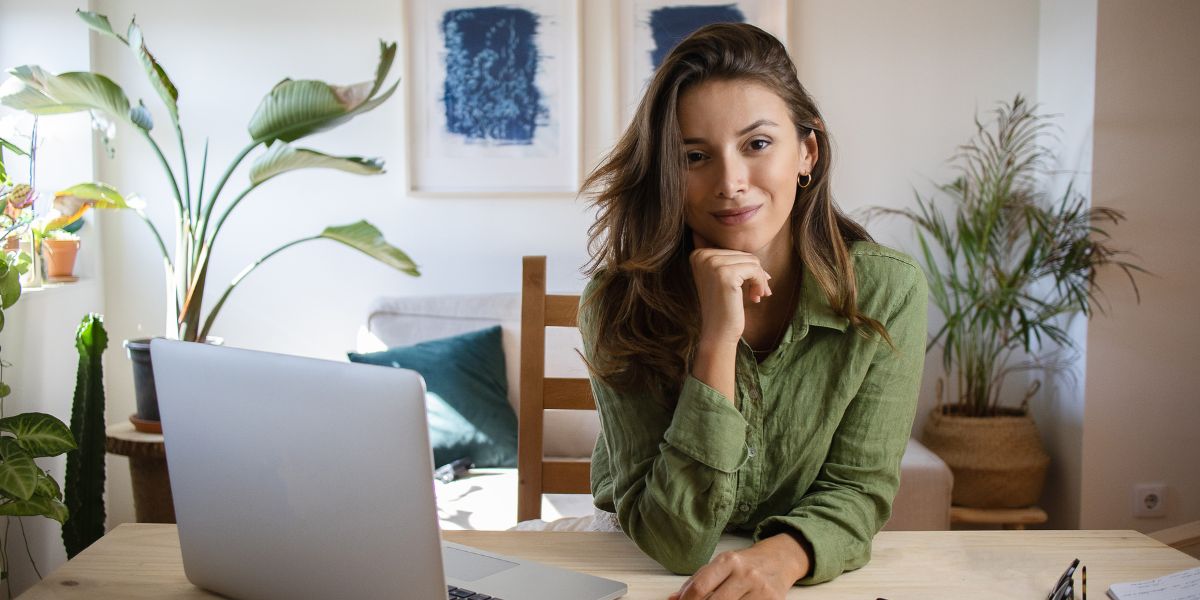 Eine junge Frau sitzt mit ihrem Laptop an einem Tisch und guckt freundlich