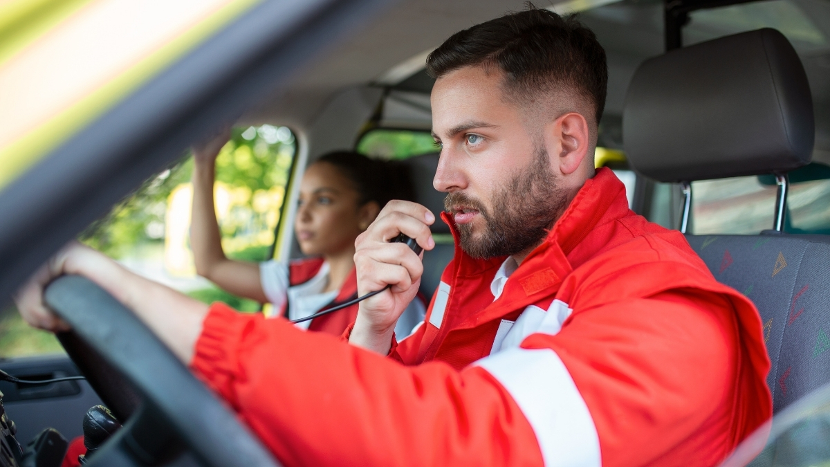 Mann in roter Einsatzkleidung spricht über Funk im Auto, während eine Beifahrerin im Hintergrund sitzt.