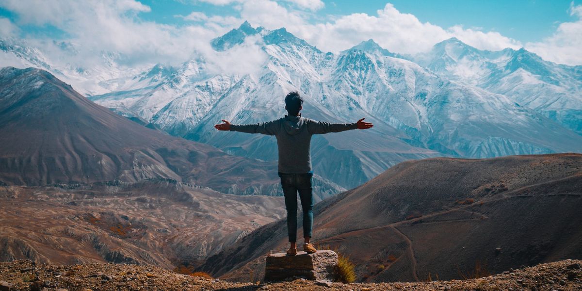 Person mit ausgebreiteten Armen steht auf einem Felsen vor einer schneebedeckten Bergkette unter blauem Himmel mit Wolken.