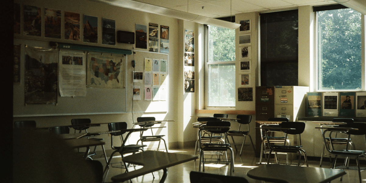 Leeres Klassenzimmer mit Tischen, Stühlen, Tafel, Unterrichtsmaterialien und großen Fenstern.
