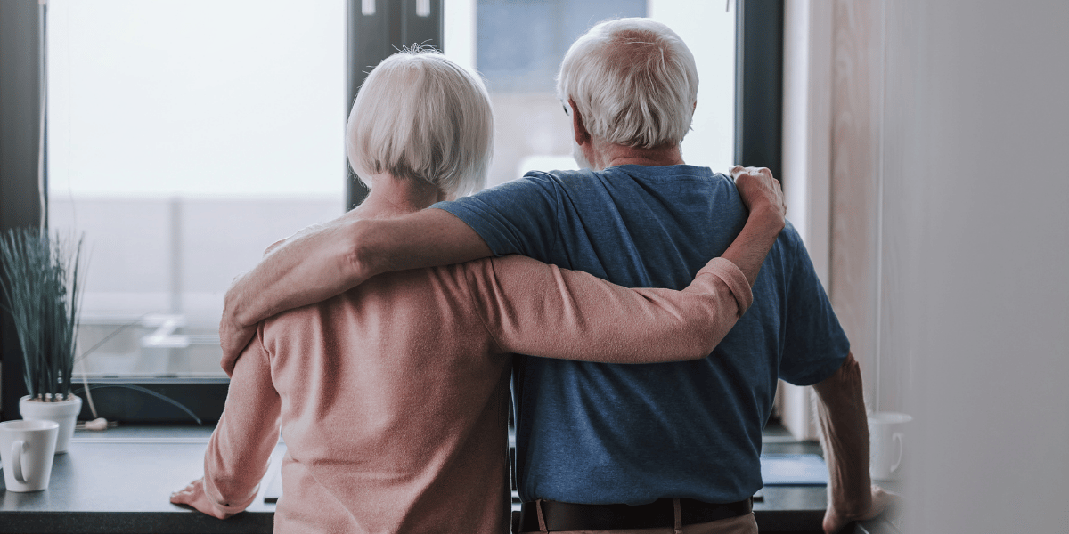 Zwei ältere Personen stehen Arm in Arm mit dem Rücken zur Kamera an einem Fenster.