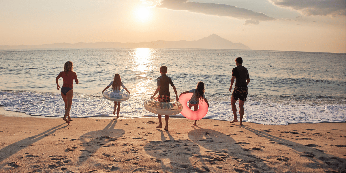 Fünf Personen laufen mit Schwimmringen am Sandstrand ins Meer, die Sonne steht tief über dem Horizont.