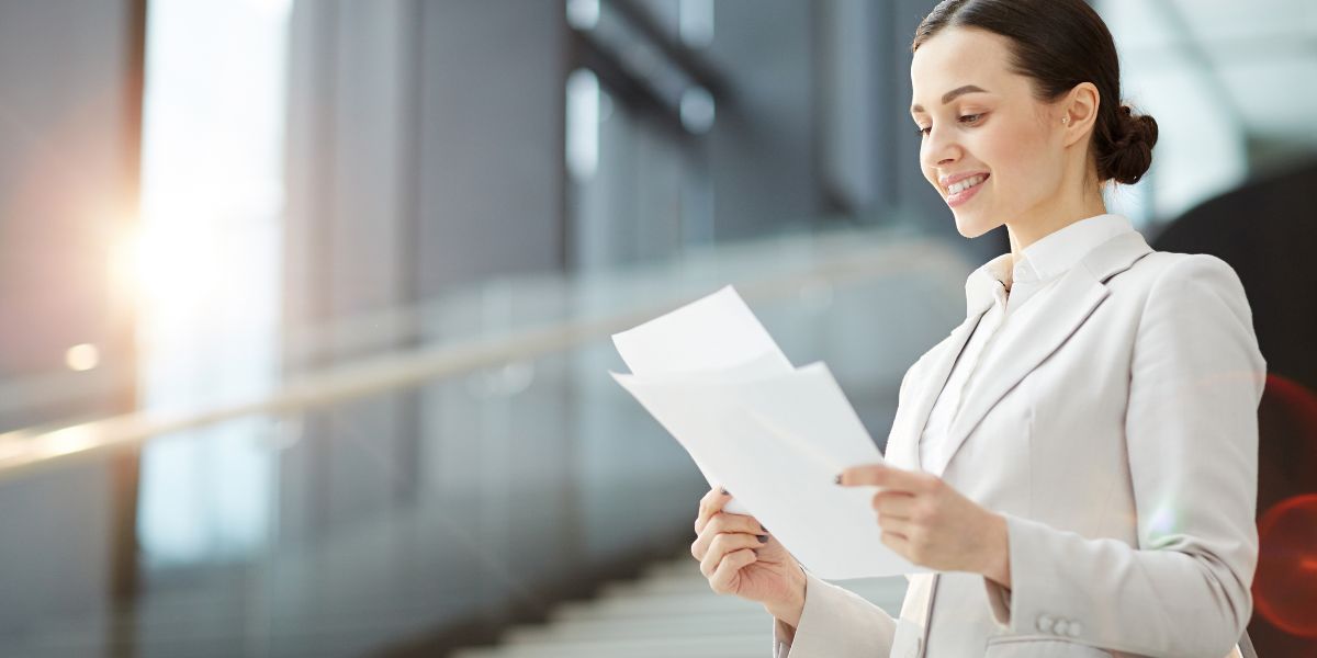L&auml;chelnde Frau im Business-Outfit liest Dokumente in einem modernen B&uuml;ro.