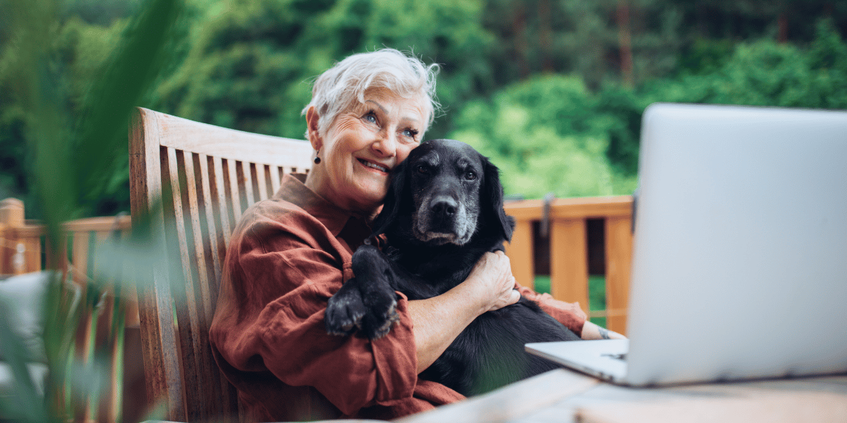 Ältere Person sitzt mit einem schwarzen Hund im Arm an einem Tisch vor einem Laptop im Garten.