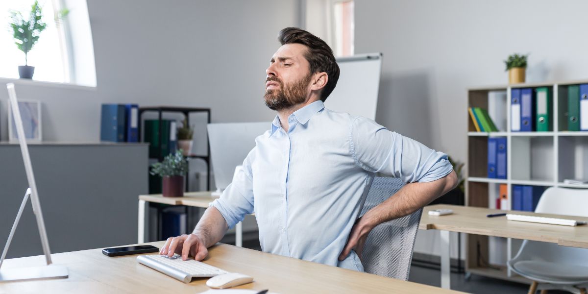 Ein Mann sitzt mit Rückenschmerzen im Büro und guckt schmerzverzerrt