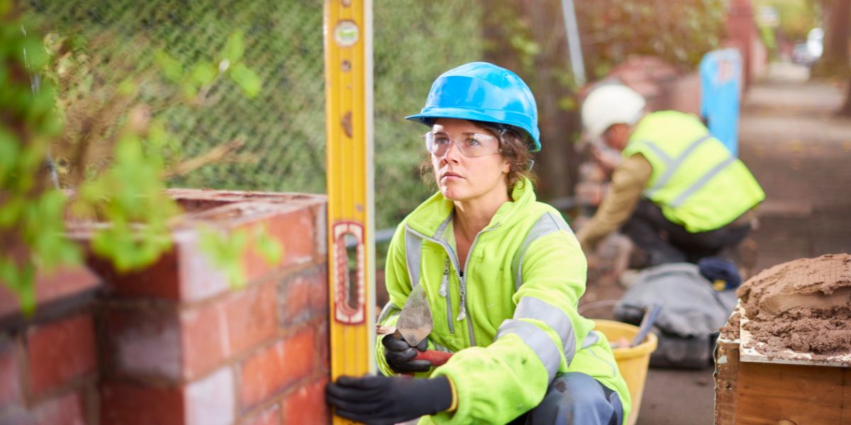 Eine Bauarbeiterin mit blauem Schutzhelm und Warnkleidung arbeitet mit einer Wasserwaage an einer Mauer; im Hintergrund sind weitere Bauleute zu sehen.