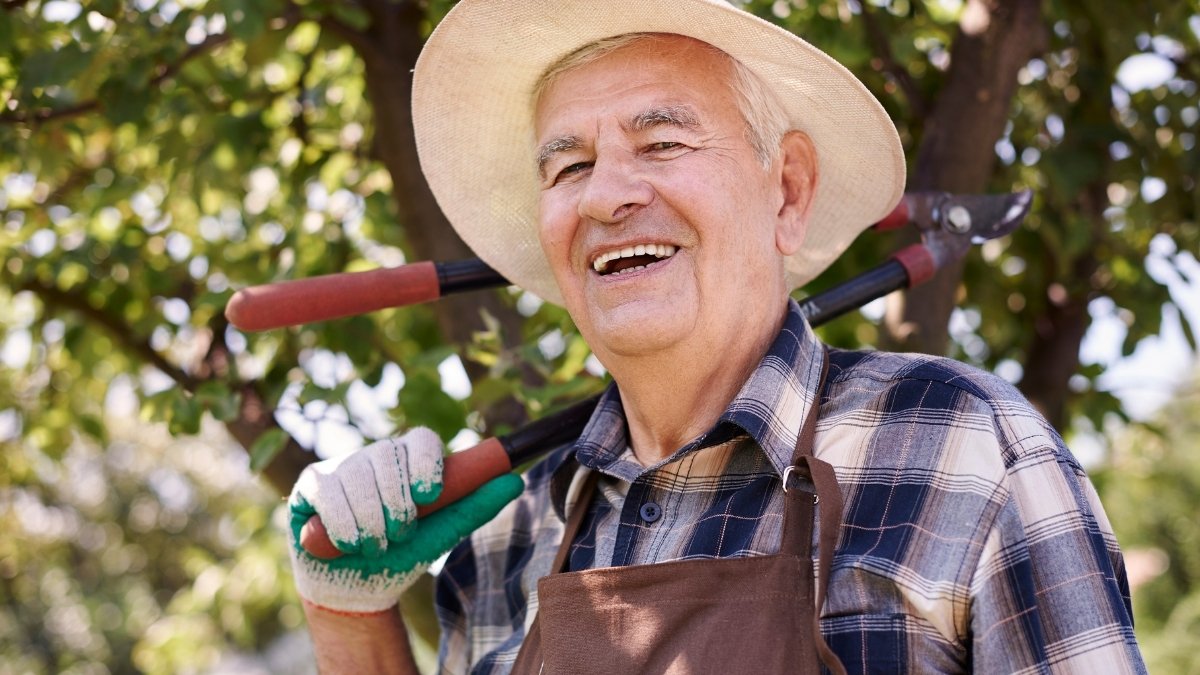 Älterer Mann mit Hut und Gartenwerkzeug steht lächelnd im Freien.