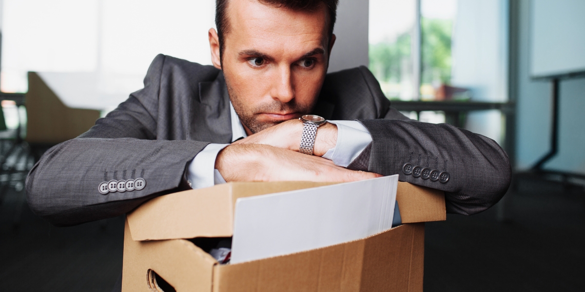 Mann im Anzug sitzt vor voller Kartonbox mit Büro im Hintergrund.